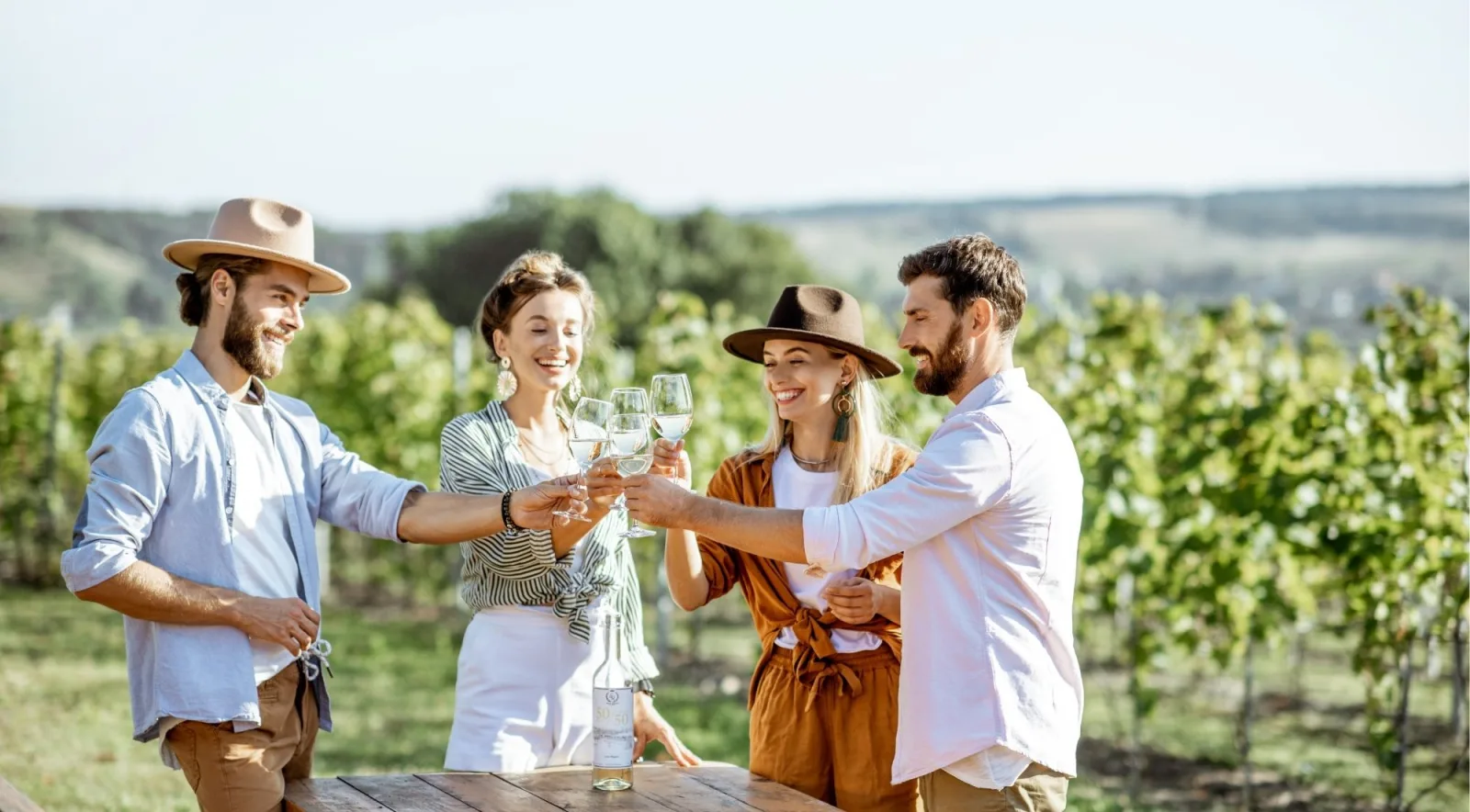 Four travellers raising glasses of white wine at a wooden table beside the vines
