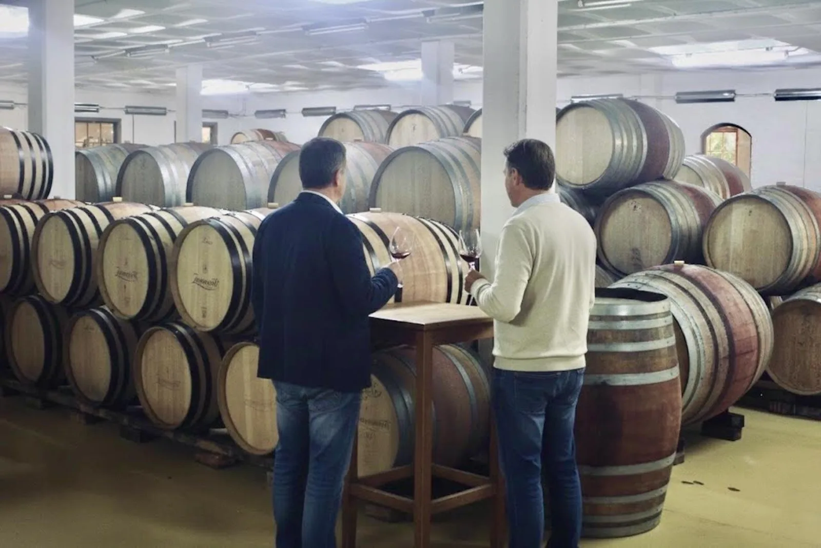 Two visitors tasting at a table among rows of oak barrels in an estate cellar