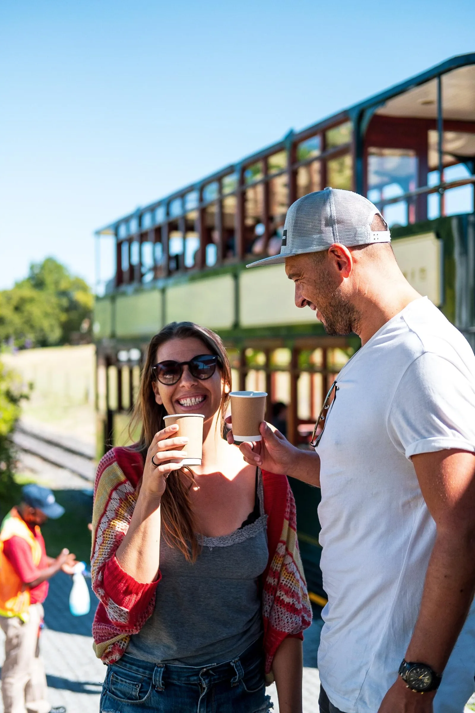 Two guests with drinks beside an open-sided tram-bus at a Franschhoek estate stop
