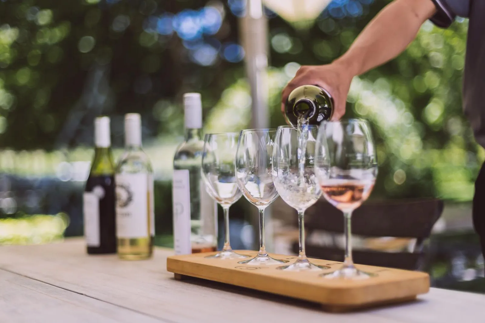A hand pouring rosé along a row of glasses on a tasting paddle beside the bottles