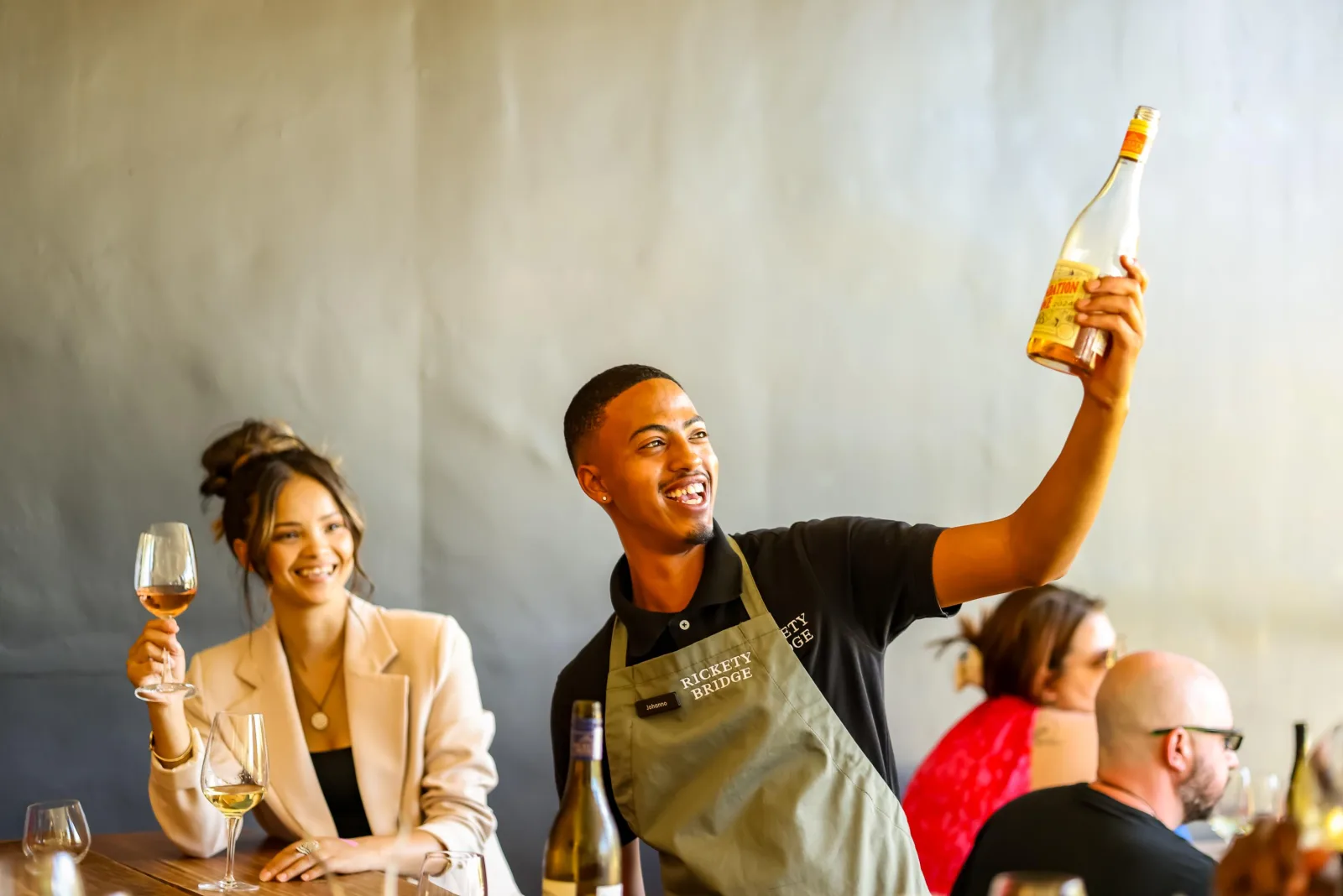 A host raising a bottle beside seated guests at an outdoor tasting